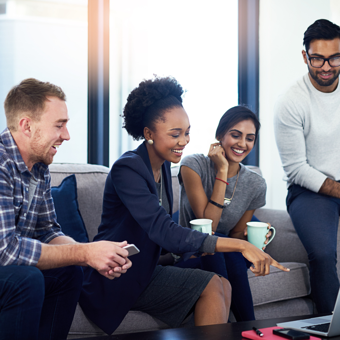 4 people in multicultural setting smiling and sitting on a couch looking at a computer screen